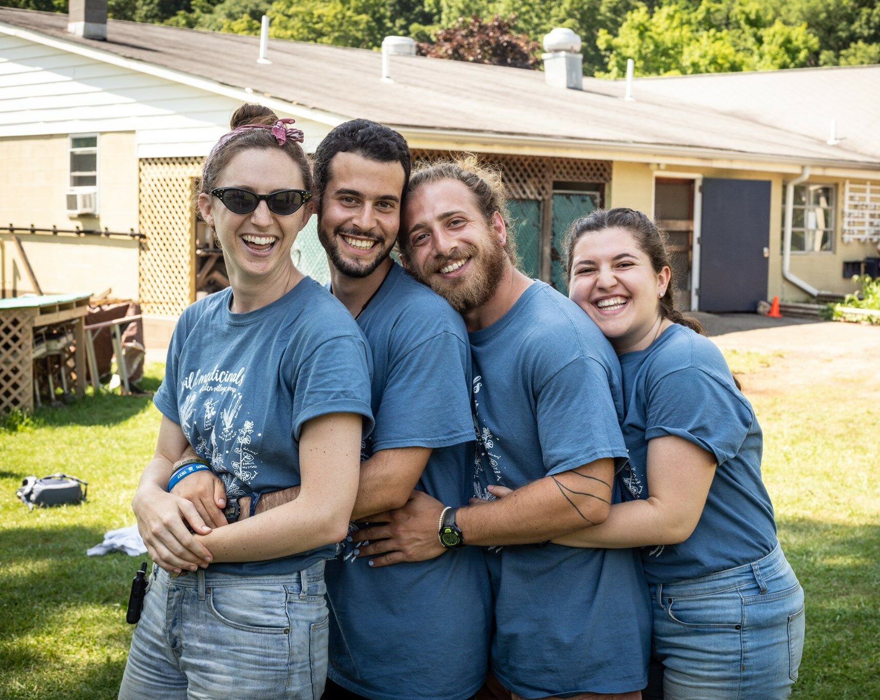 Four individuals in matching blue t-shirts smiling together outdoors near a building.