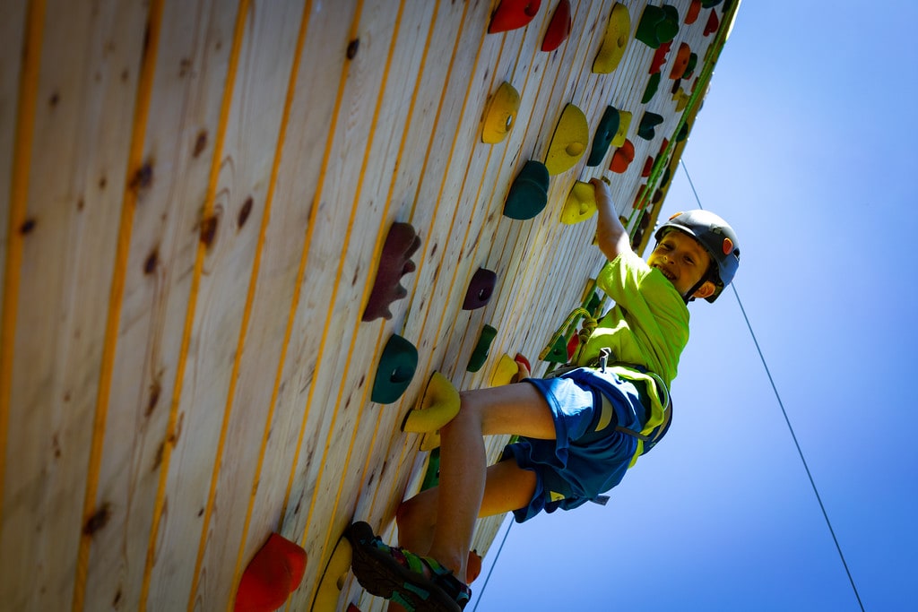 Child climbing a colorful outdoor rock wall, smiling, under a clear blue sky.