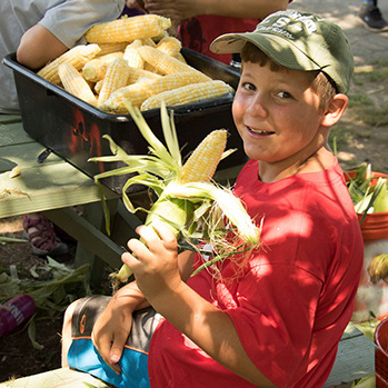 Child smiling while holding freshly harvested corn at a wooden table.