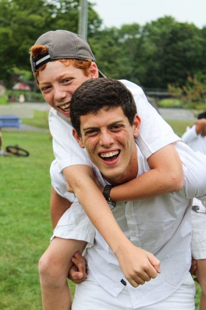 Staff member giving a camper a piggyback ride, both smiling outdoors.